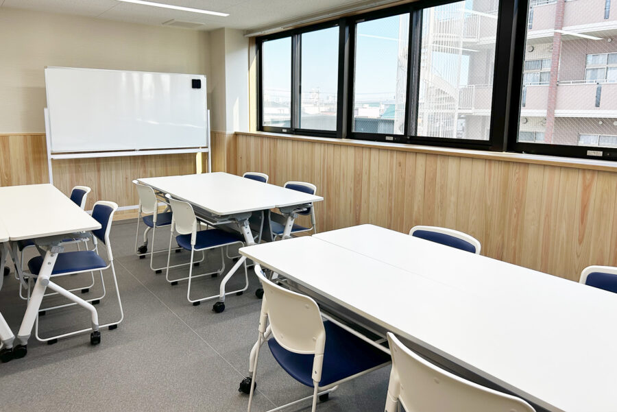 明るい共用スペース。複数のテーブルと椅子が配置され、大きな窓から採光を得ています。